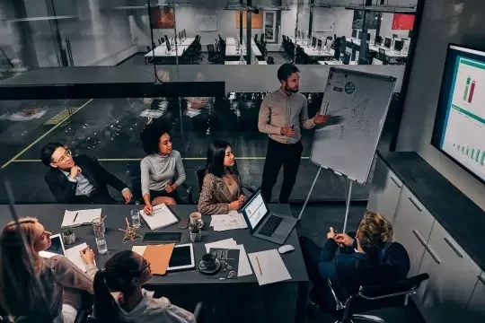 Six people sit at a meeting table in an office environment, looking towards a TV on the adjacent wall as a man stands next to a white board and explains business figures.