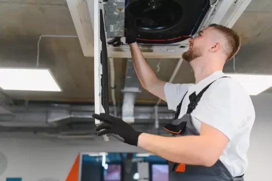An engineer fixes air ducting on a ceiling in a facility.
