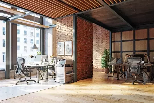A spacious office environment with three desks and monitors visible. The walls are bare brick and wood, with large windows letting in daylight.