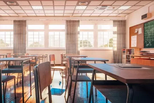 An empty, clean school classroom with desks and a blackboard. Light pours in through the windows, illuminating the wooden floor and desks.