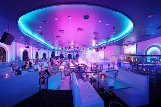 A vast club with tables. Lighting is purple and blue. A few people stand at a bar to the right but the venue is mostly empty.