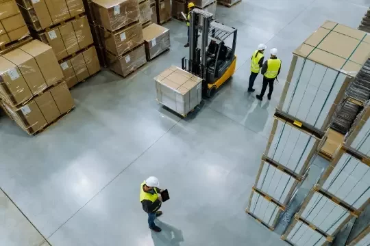 Organised pathway through a warehouse where a forklift can pass through. Multiple people in high-vis stand at the side.